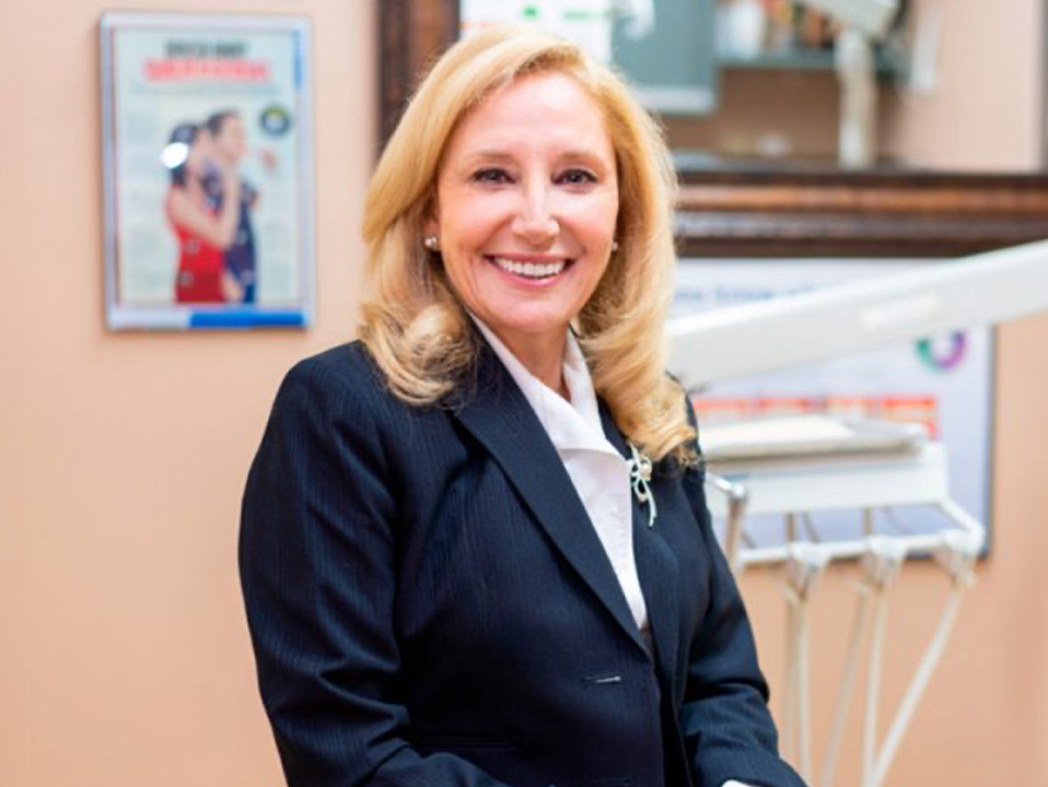 The image shows a woman standing in front of a dental office setting, wearing a black blazer, smiling at the camera with her hands clasped together.