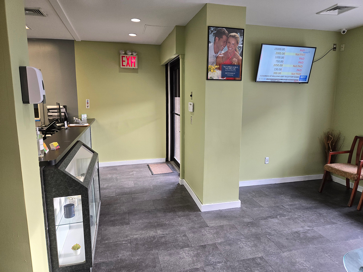 The image shows an interior view of a restaurant or cafe with a clean and well-maintained appearance, featuring green walls, a light gray floor, a counter area with a cash register, a menu board, and a TV screen displaying a price list, as well as a framed picture on the wall.